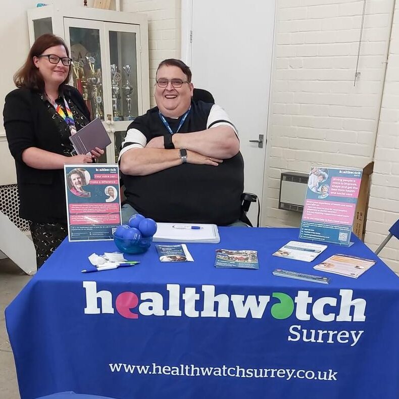 Photo of Shaun at an Age UK event, Shaun is seated in his wheelchair, behind a table branded with Healthwatch Surrey tablecloth, merchandise and leaflets